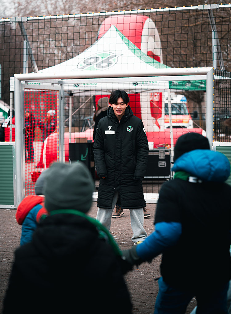 Hayate Matsuda steht dick angezogen im Tor, vor ihm unscharf drei Kinder.