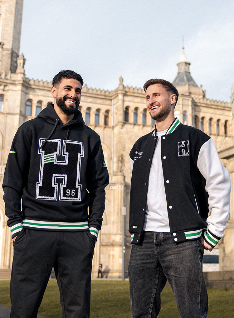 Enzo Leopold in einer Collegejacke und Elias Saad in einem Hoodie stehen vor einem großen alten Gebäude.