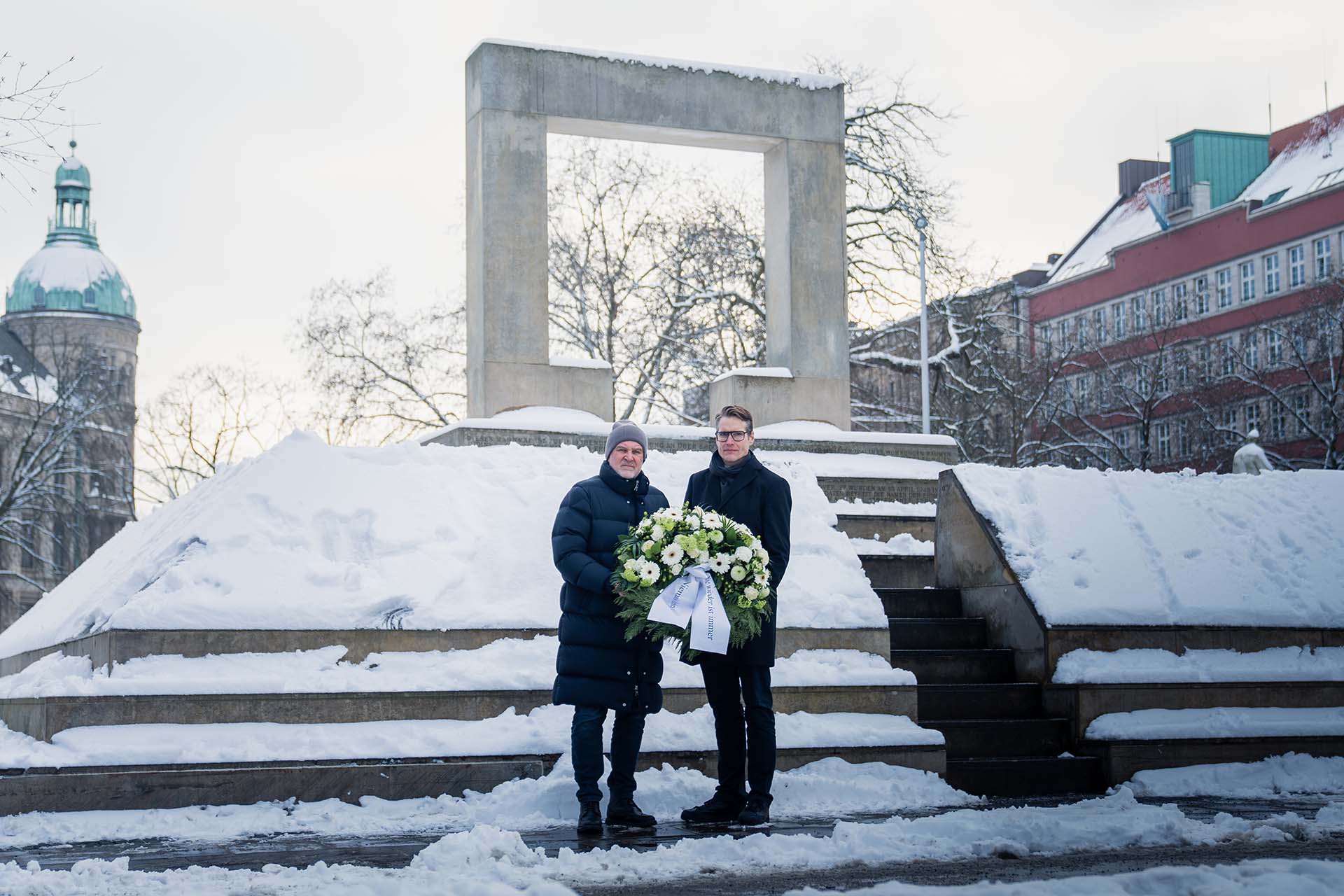 Jörg Schmadtke und Henning Bindzus stehen dick angezogen vor dem Holocaust-Mahnmal und halten gemeinsam einen Kranz. Es liegt Schnee.