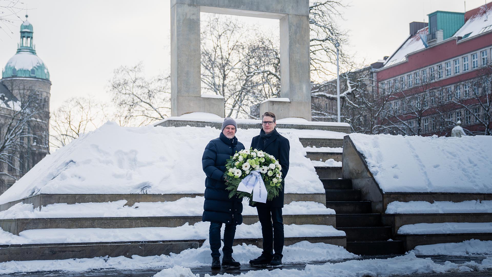 Jörg Schmadtke und Henning Bindzus stehen dick angezogen vor dem Holocaust-Mahnmal und halten gemeinsam einen Kranz. Es liegt Schnee.