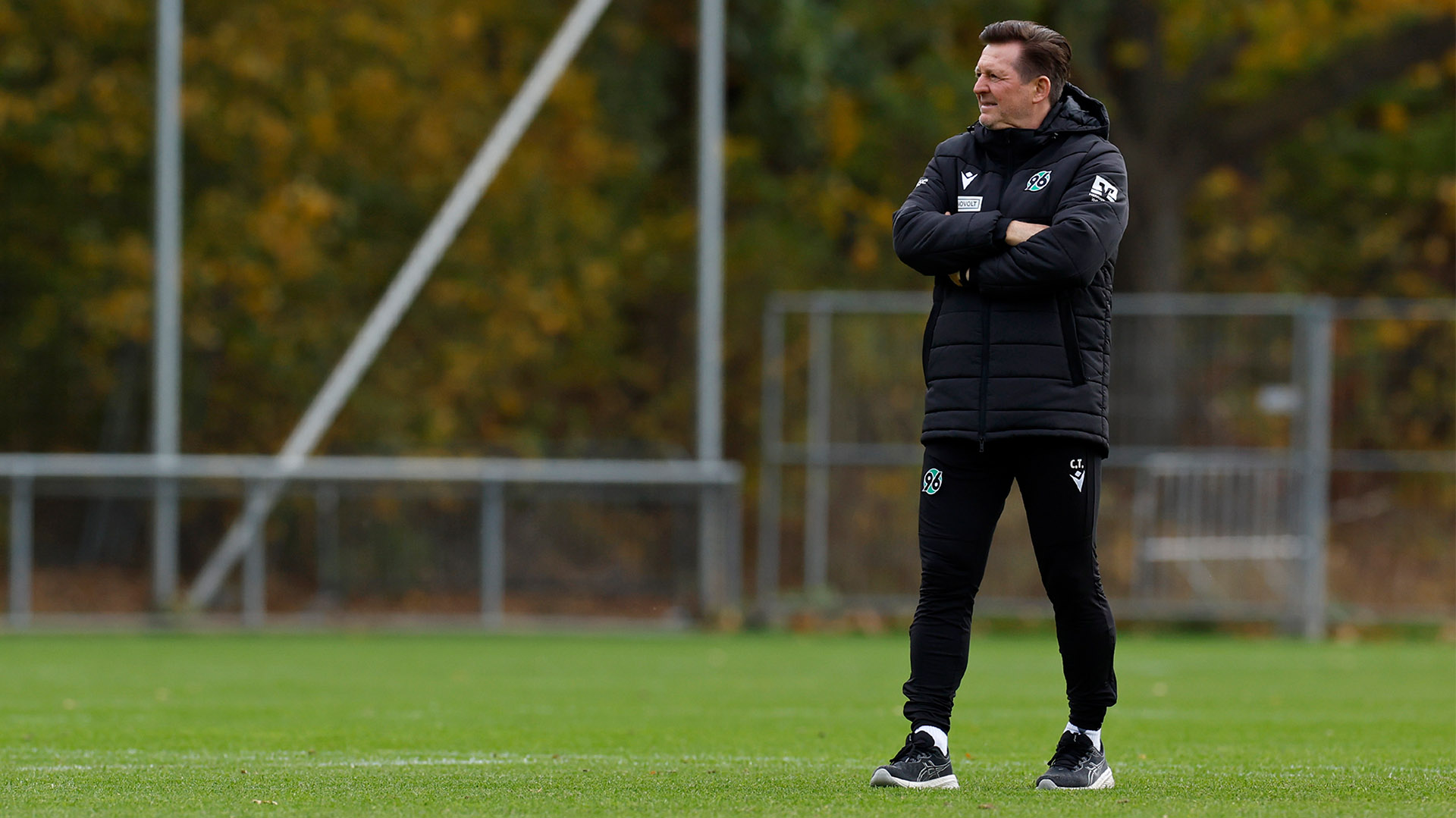 Ganzkörperaufnahme von Christian Titz auf dem Trainingsplatz, er verschränkt die Arme und schaut nach rechts.