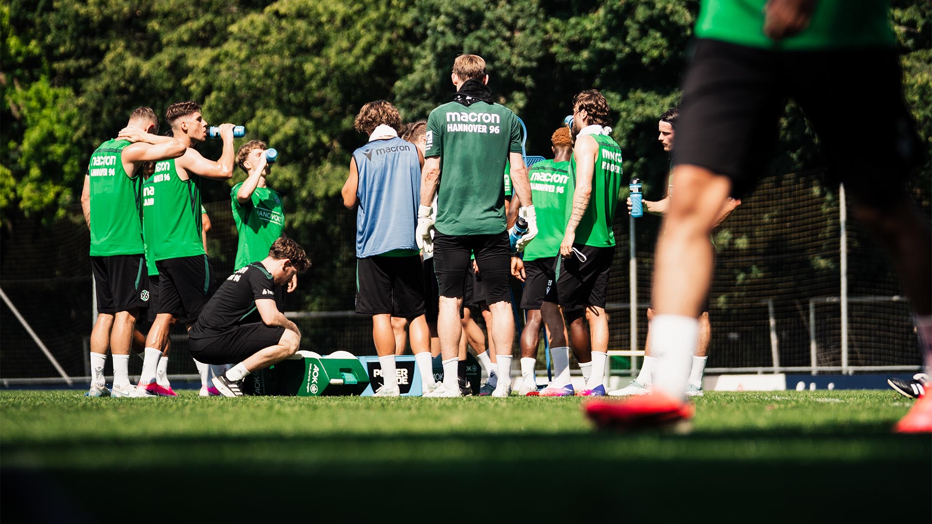 Ein Gruppe Spieler steht im Kreis auf dem Trainingsplatz und pausiert, einige trinken etwas. 