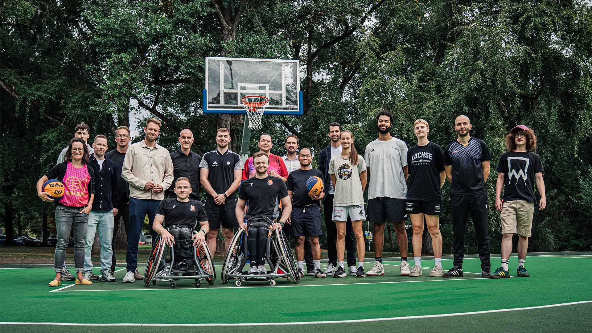 Eine Gruppe von rund 20 Männern und Frauen steht auf einem grünen Sportplatz mit einem Basketballkorb im Hintergrund. 