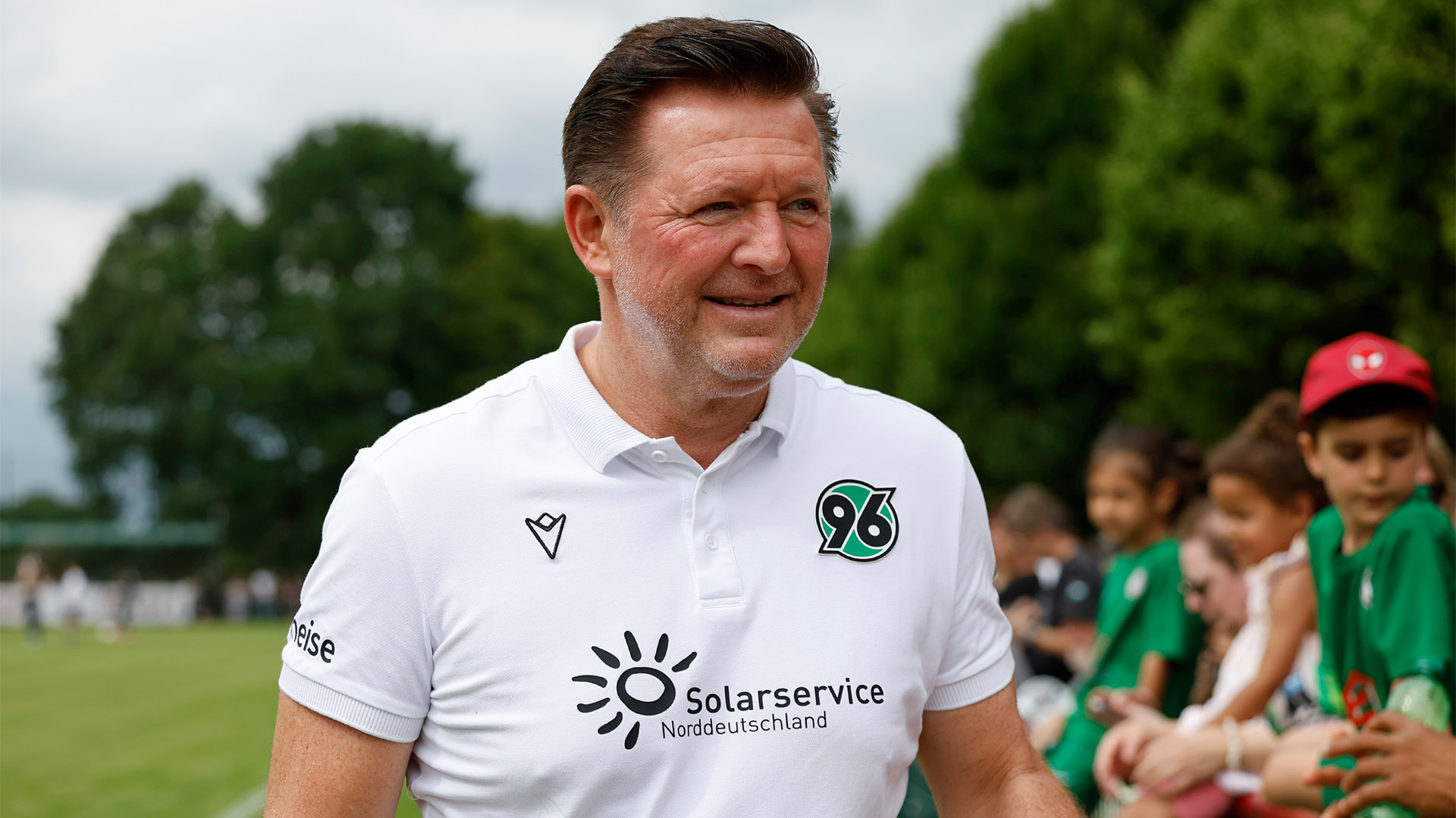 Hannover-96-Coach Christian Titz lächelt am Seitenrand beim Test gegen den HSC, im Hintergrund sind unscharf Fans zu sehen. 