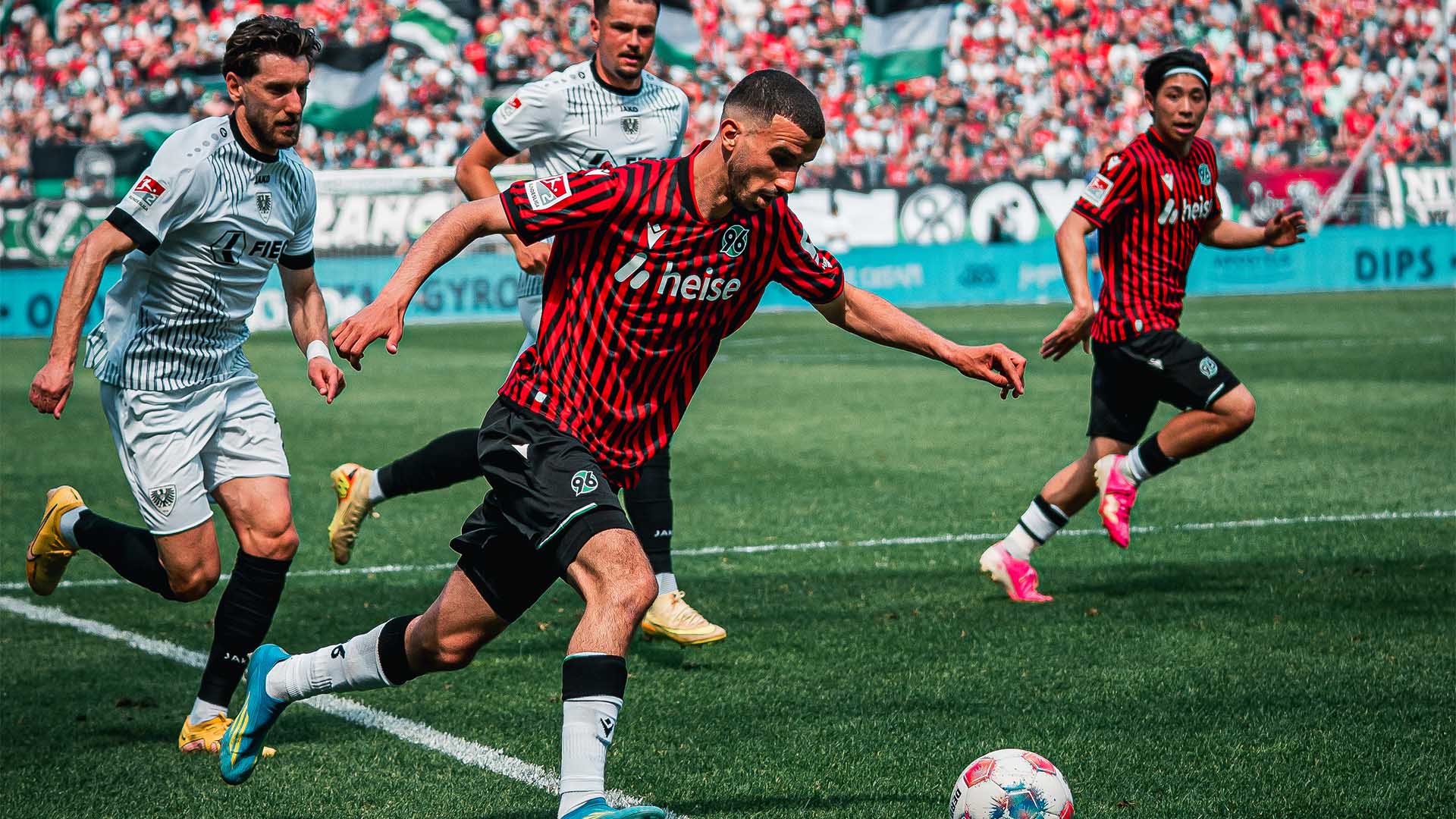 Waniss Taibi im Laufen am Ball, im Hintergrund zwei Münster- und ein 96-Spieler sowie die Fans auf den Rängen.