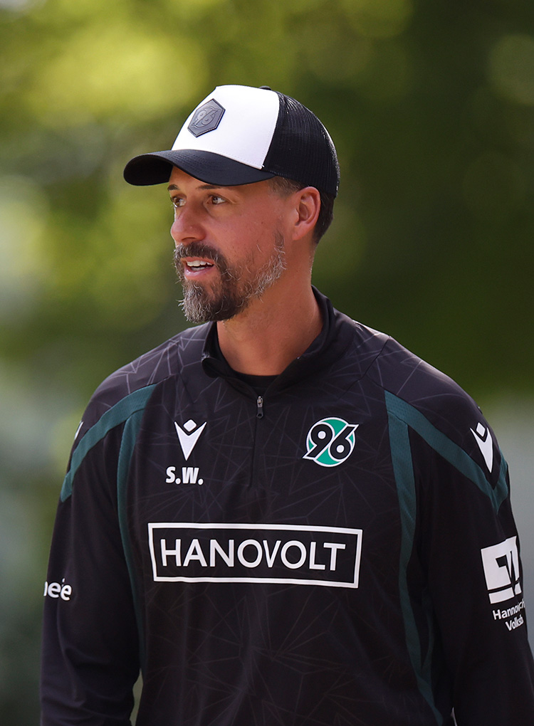 96-Cheftrainer Sandro Wagner in Trainingskleidung und mit Cap ist bis zur Brust zu sehen, er schaut leicht nach rechts.