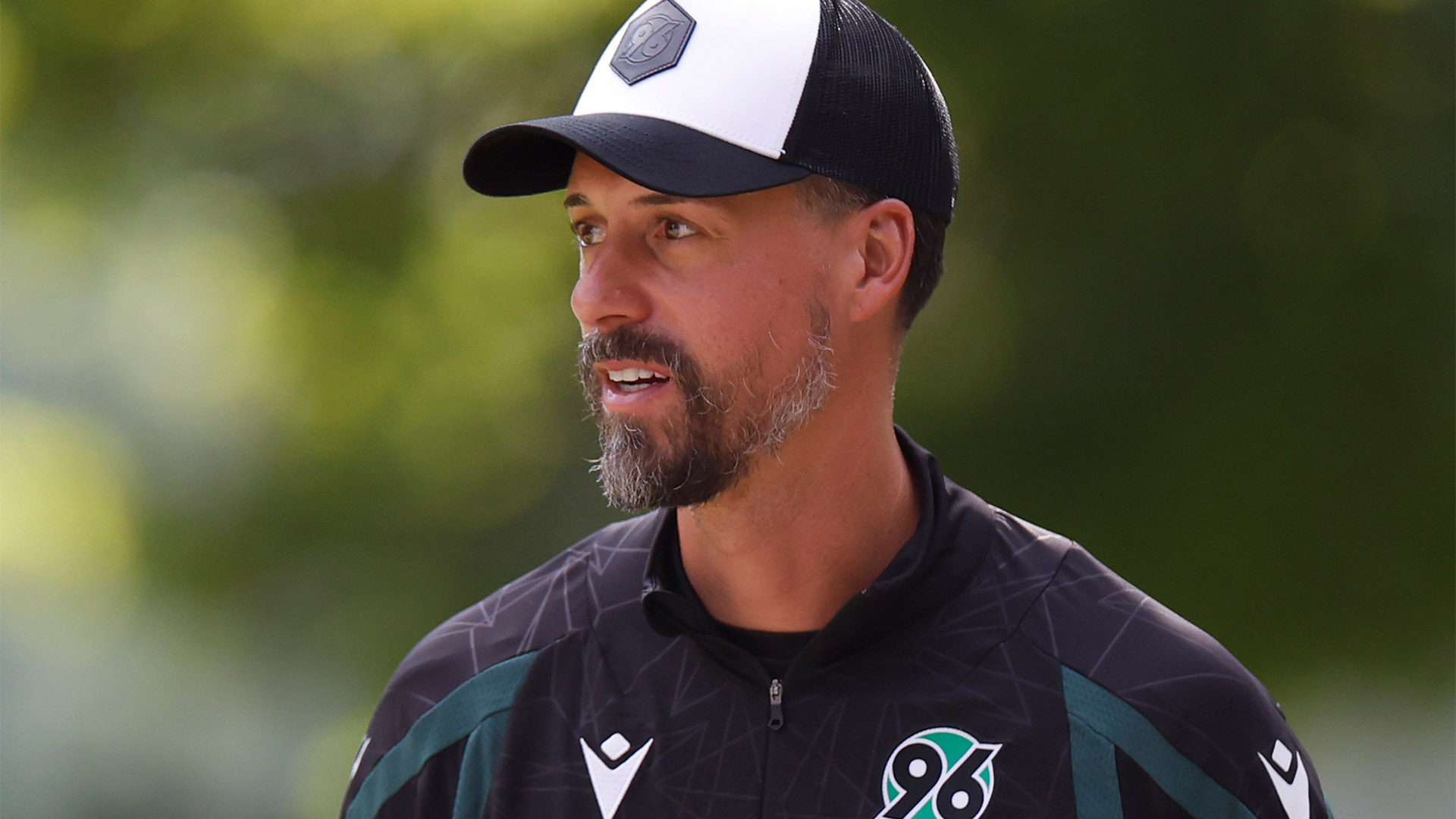 96-Cheftrainer Sandro Wagner in Trainingskleidung und mit Cap ist bis zur Brust zu sehen, er schaut leicht nach rechts.