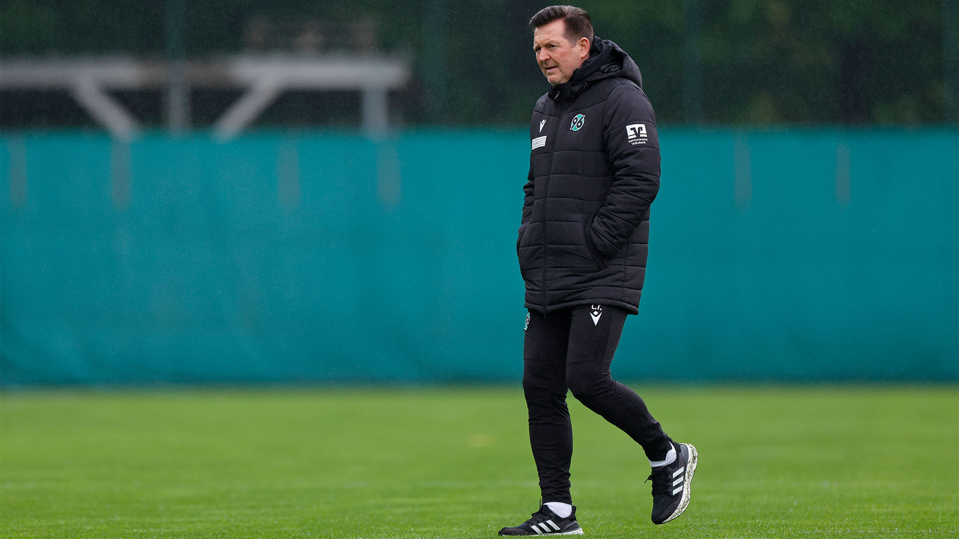 Christian Titz läuft mit Händen in den Taschen in Winterjacke und langer Trainingshose über den Platz.