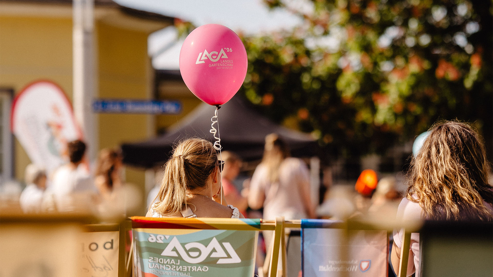 Menschen im Liegestuhl von hinten, eine Person hält einen rosafarbenen Luftaballon mit der Aufschrift "Laga '26".