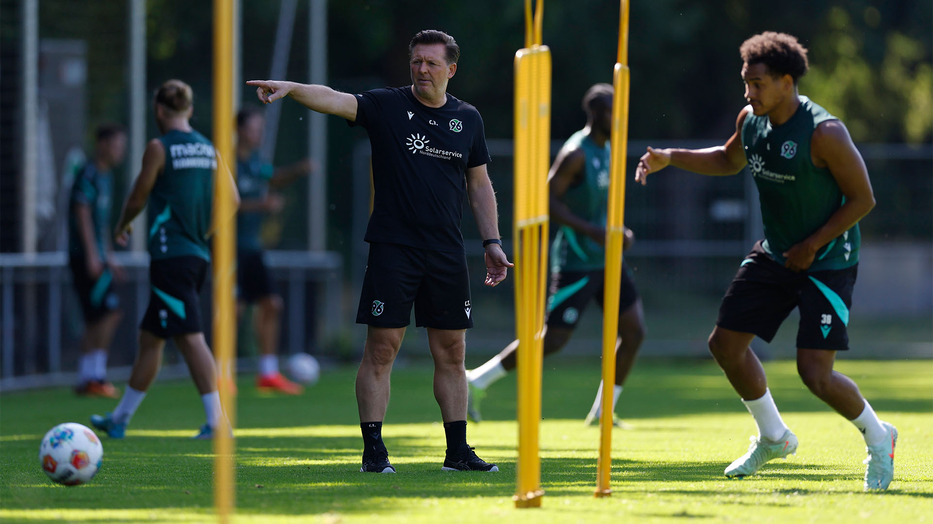 Hannover-96-Trainer Christian Titz steht auf dem Trainingplatz und zeigt mit dem rechten Arm nach rechts.