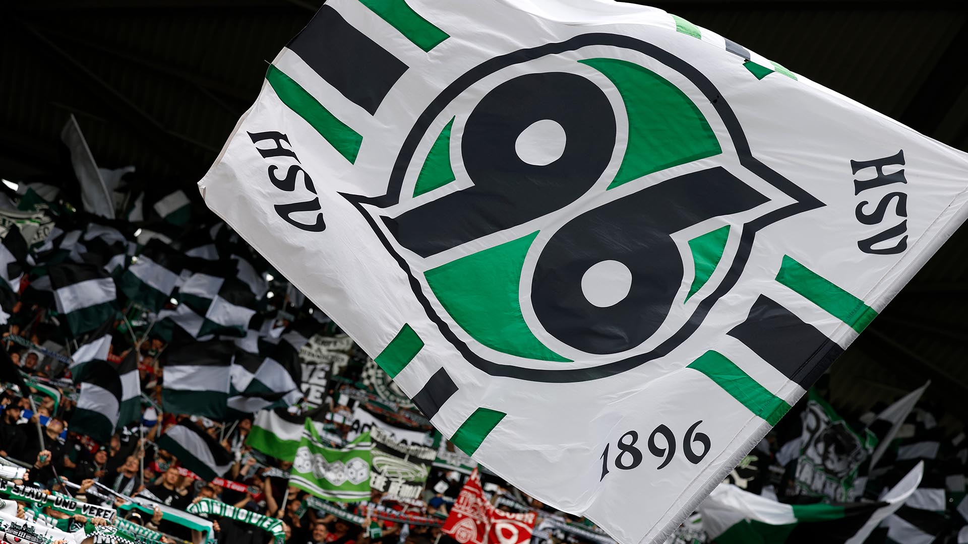 Eine große 96-Fahne weht vor der Nordkurve des Stadions, im Hintergrund sind viele Schals und weitere Fahnen zu sehen.