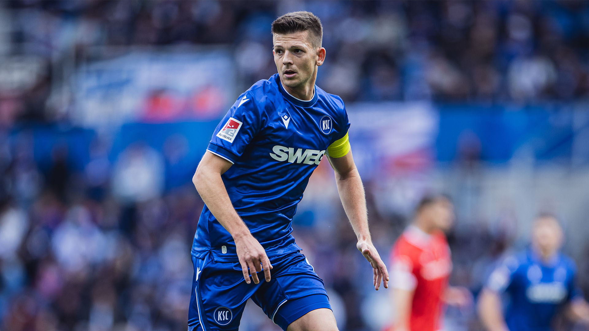 Marvin Wanitzek läuft im blauen Trikot über das Spielfeld. Er trägt eine gelbe Kapitänsbinde. Im Hintergrund ist ein unscharfes Stadion voller Zuschauer zu sehen. 