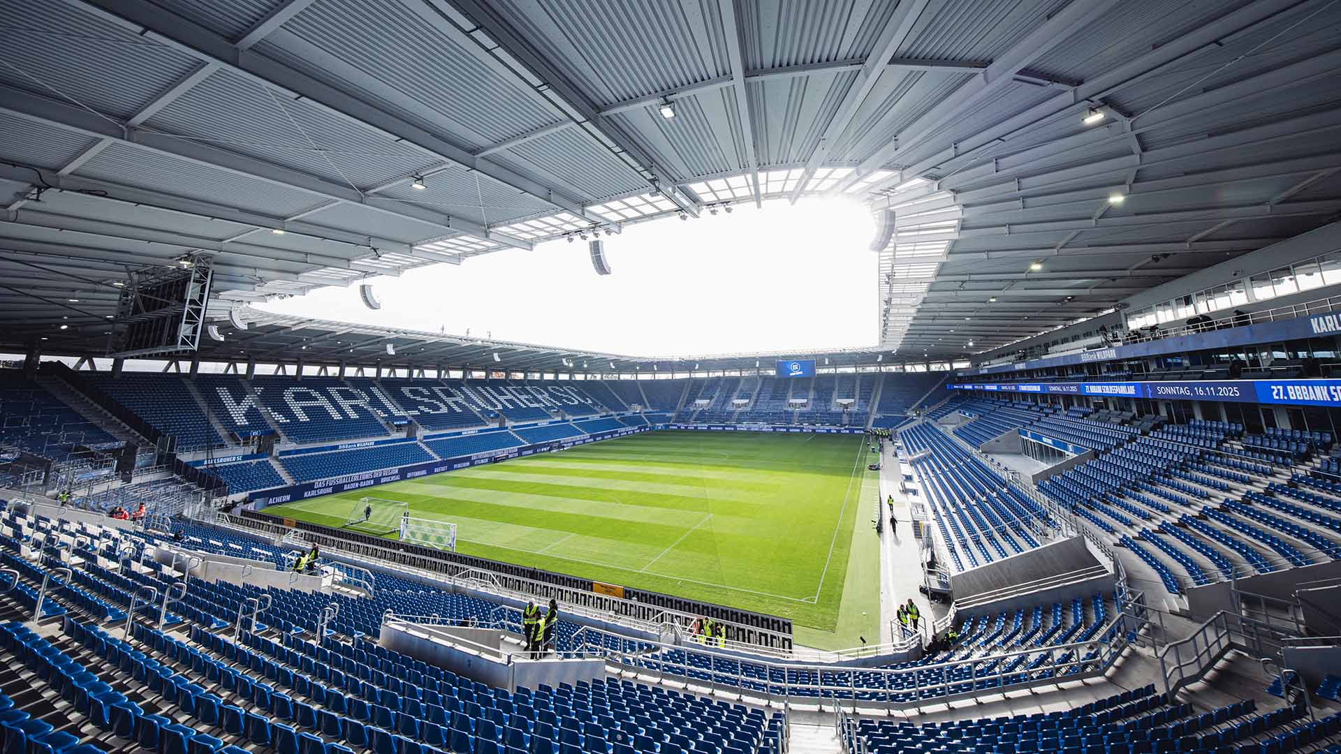 Leeres Stadion des Karlsruher SC, aufgenommen aus einer Ecke der Tribüne, mit überwiegend blauen Sitzen; weiße Sitze bilden den Vereinsnamen auf einer der Geraden.