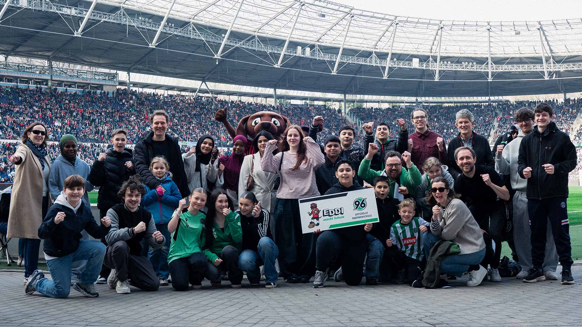 Eine große Gruppe von Kindern, Jugendlichen, Erwachsenen und Maskottchen EDDI posiert gemeinsam im Innenraum der Heinz von Heiden Arena. Viele heben jubelnd die Fäuste, während sie eine Plakette mit der Aufschrift „EDDI macht Schule“ in die Kamera halten.