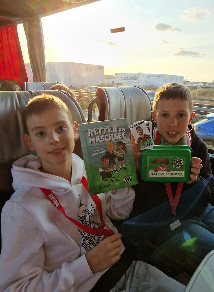 Zwei Kinder sitzen im Bus und halten das Buch "Retter am Maschsee", eine Brotbüchse mit EDDI-Aufdruck und 96-Spielkarten in die Kamera. Im Hintergrund ist der Sonnenuntergang zu sehen.