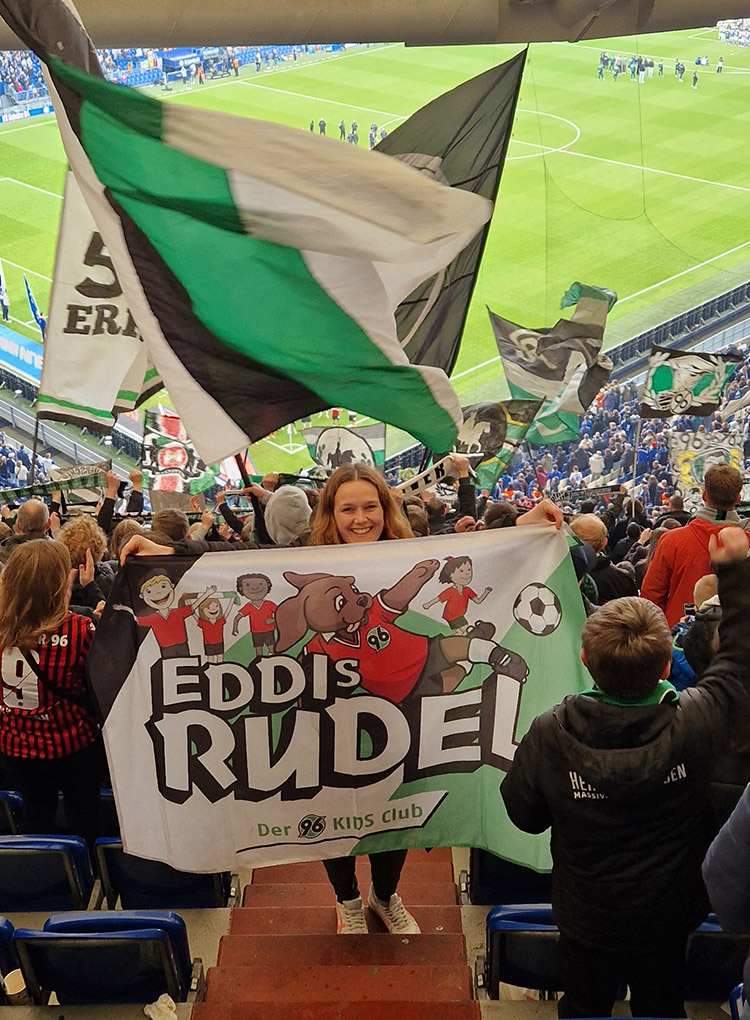 Die Fahne "EDDIs Rudel" wird auf einer Treppe in der Fankurve hochgehalten. Im Hintergrund sind Fahnen und das Spielfeld zu sehen.