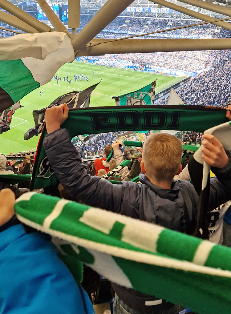 Kinder stehen mit dem Rücken zur Kamera, schauen in Richtung Spielfeld und halten Schals von Hannover 96 in die Luft.