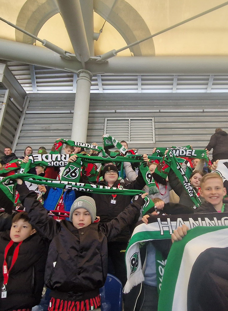 Kinder stehen in der Fankurve, schauen in die Kamera und halten Schals von Hannover 96 in die Luft.