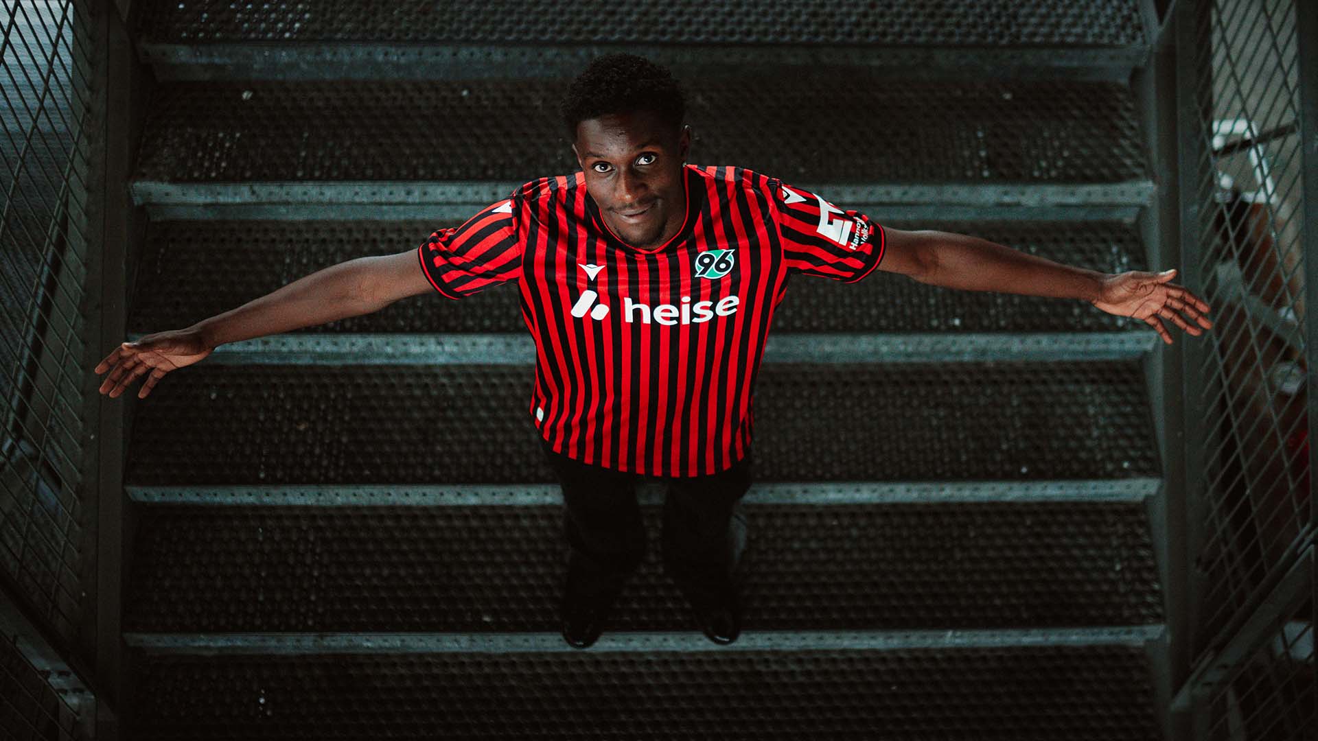 William Kokolo steht im Heimtrikot auf einer Treppe im Stadion, schaut von unten in die Kamera und breitet beide Arme aus.
