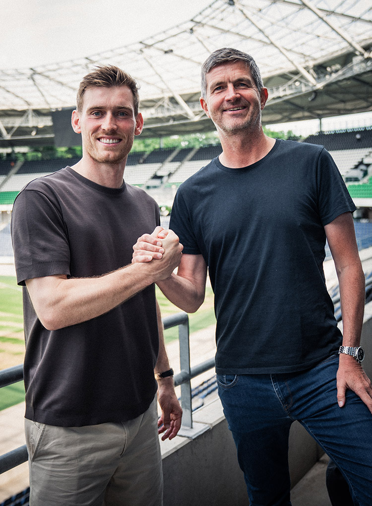 Leo Weinkauf und Ralf Becker beim Handshake im Stadion, dahinter die schwarz-weiß-grüne Nordtribüne.