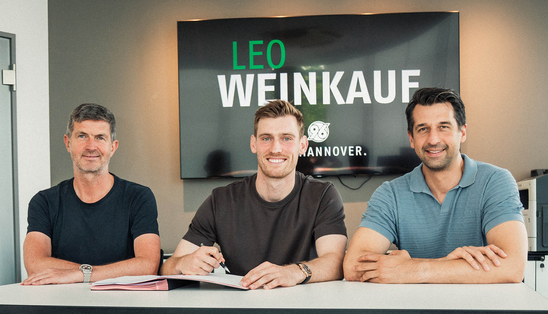 Ralf Becker, Leo Weinkauf und Jonas Boldt sitzen im Büro am Tisch, dahinter ein Screen mit der Aufschrift "Leo Weinkauf".