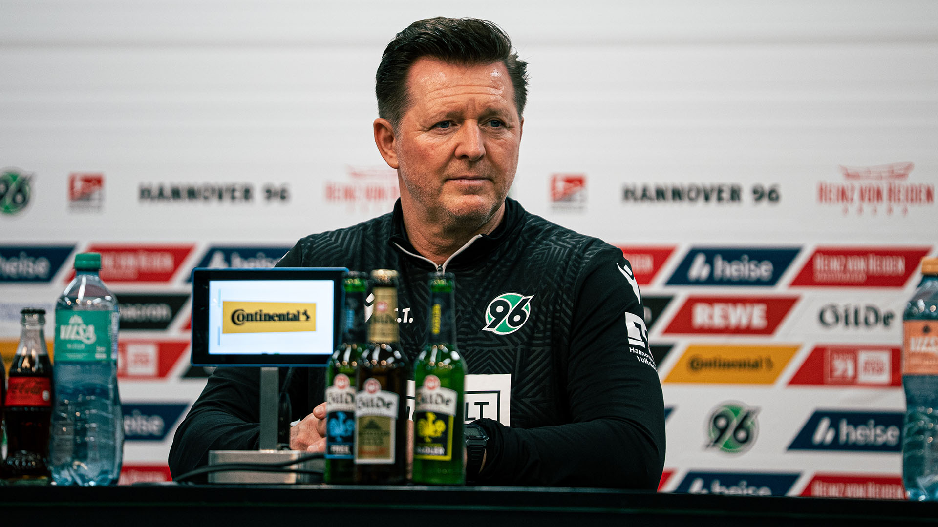 Christian Titz sitzt auf dem PK-Podium, vor ihm Flaschen und ein Tablet, im Hintergrund die 96-Sponsorenwand.
