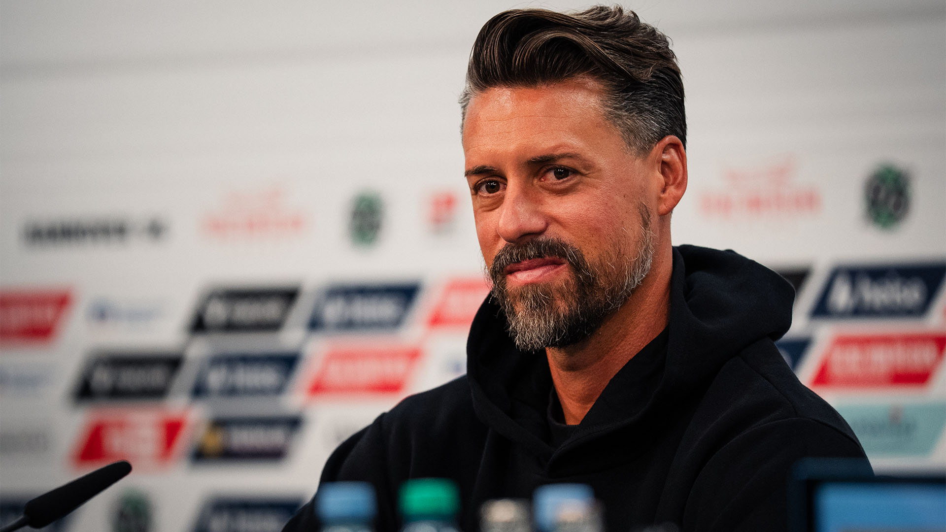 Der 96-Cheftrainer Sandro Wagner sitzt auf dem Pressekonferenz-Podium, hinter ihm unscharf eine Sponsorenwand.