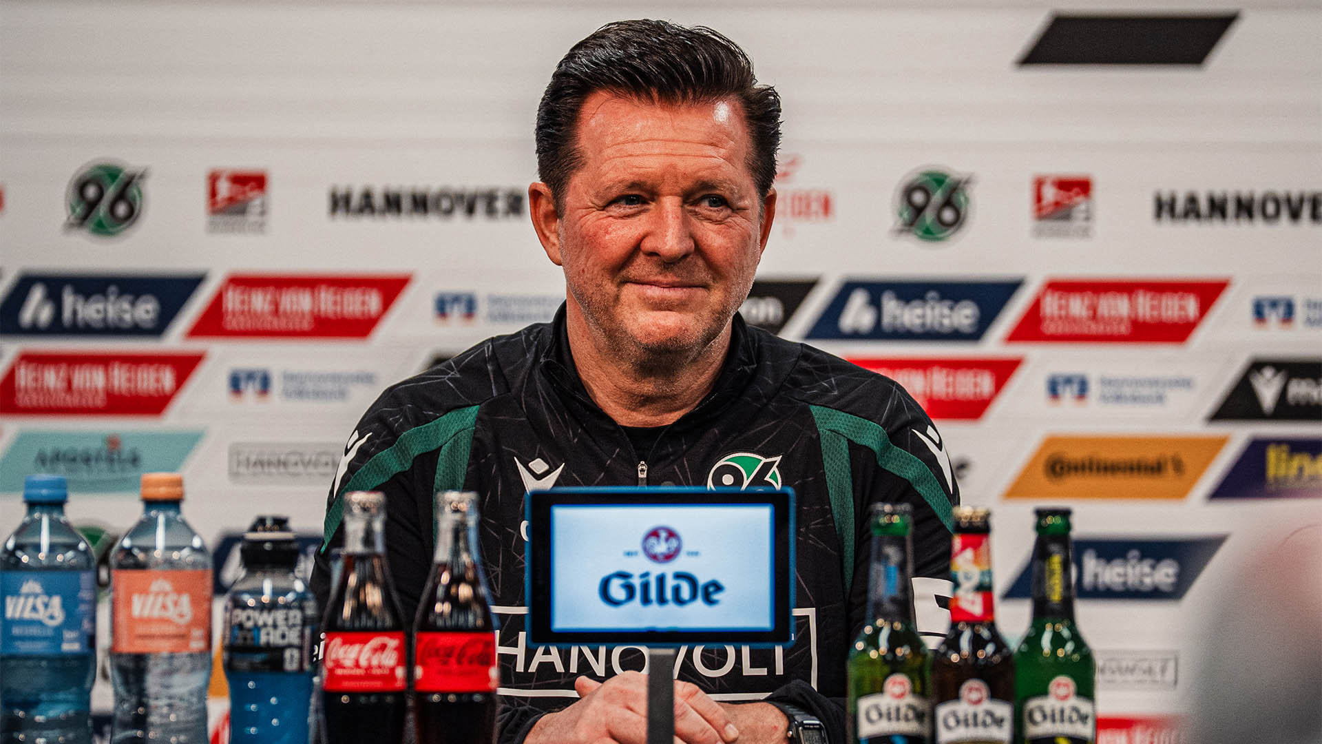 96-Cheftrainer Christian Titz sitzt auf dem Pressekonferenz-Podium vor einer Sponsorenwand, vor ihm stehen verschiedene Getränkeflaschen.