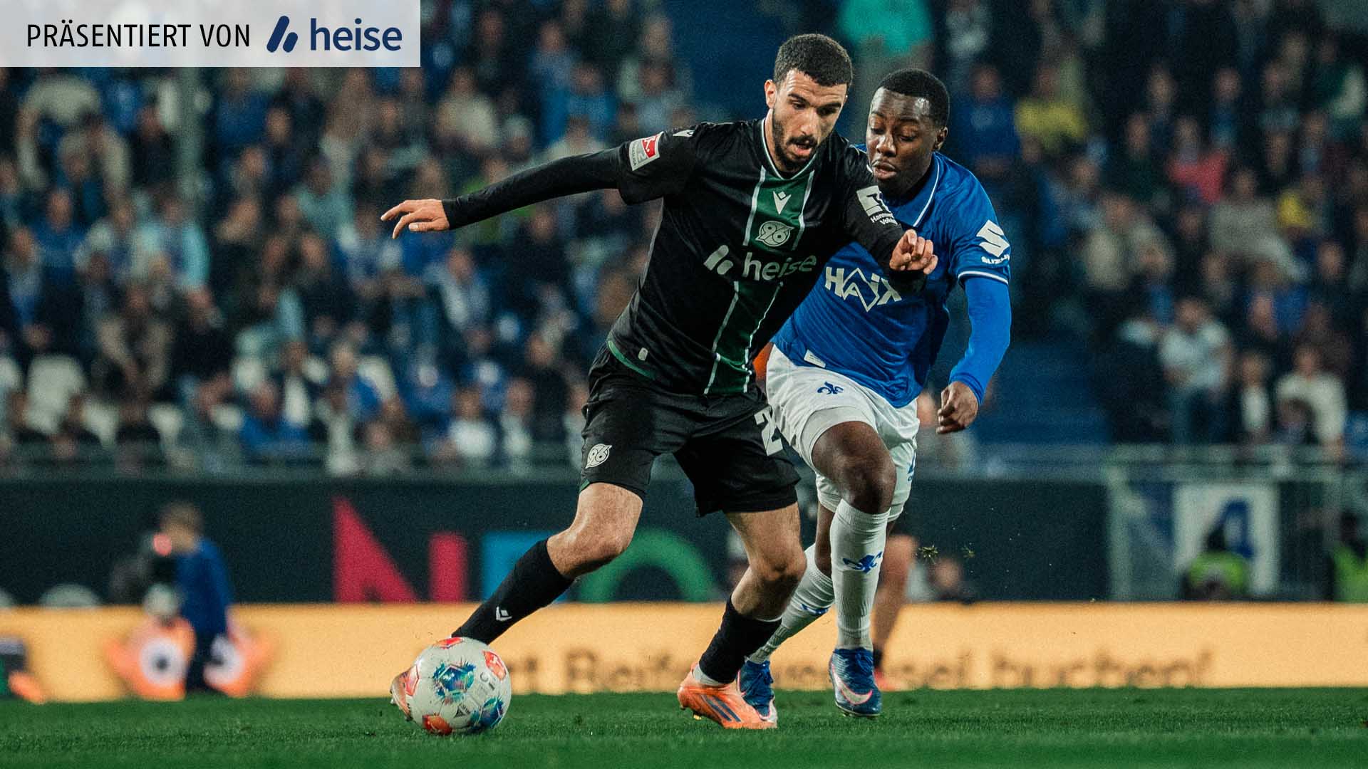 96-Spieler Waniss Taibi und Darmstadt-Spieler Merveille Papela im Duell, Taibi ist am Ball, im Hintergrund volle Ränge. 