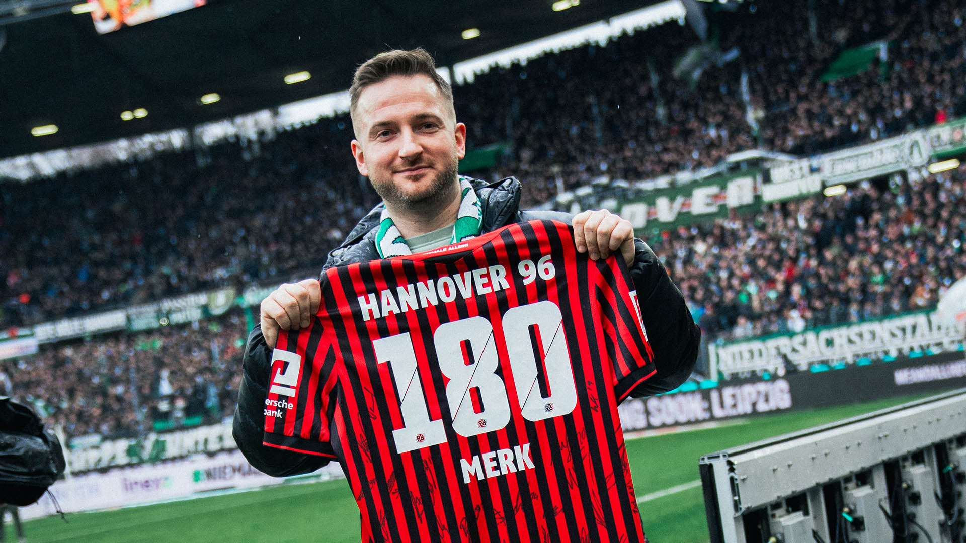 Arno Merk mit dem rot-schwarz gestreiftem Trikot in den Händen. Er zeigt die Rückseite mit der Trikotnummer in die Kamera. im Hintergrund ist das Spielfeld zu sehen.
