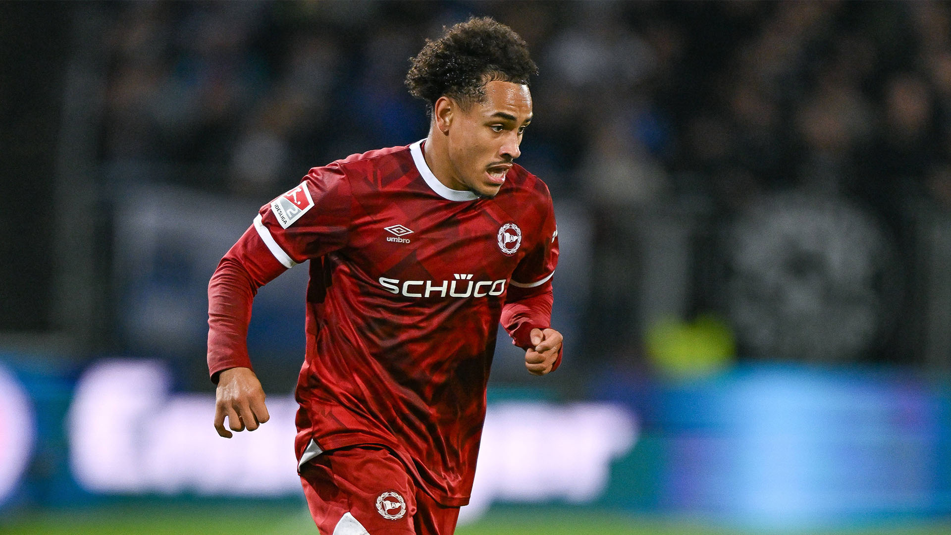 Monju Momuluh läuft über den Rasen. Er trägt ein rotes Trikot mit weißen Akzenten und ein rotes Unterarmshirt.