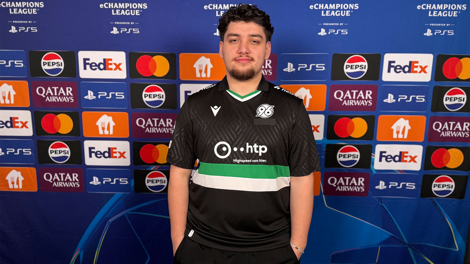 Mustafa Cankal im 96-eFootball-Trikot steht vor einer Champions-League-Sponsorenwand.