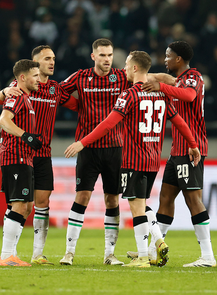 Mehrere Spieler von Hannover 96 in rot-schwarz gestreiften Heimtrikots stehen eng im Kreis zusammen auf dem Spielfeld und legen die Arme umeinander.
