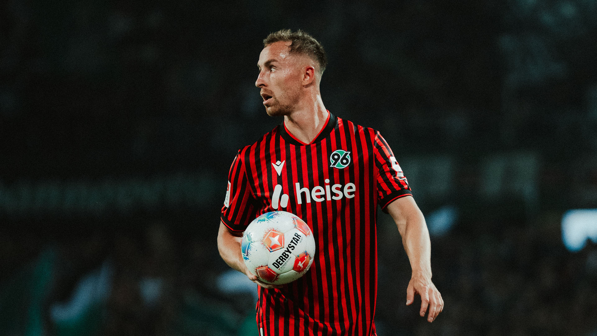 Maurice Neubauer im rot-schwarz gestreiften Heimtrikot von Hannover 96 hält einen Ball in den Händen. Er blickt konzentriert zur Seite. Das Stadion im Hintergrund ist dunkel und unscharf.