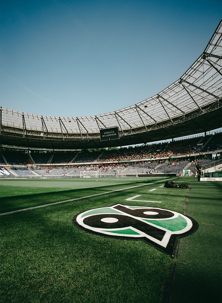 Blick auf die leicht gefüllte Nordkurve des Stadions, im Vordergrund liegt ein 96-Logo-Teppich.