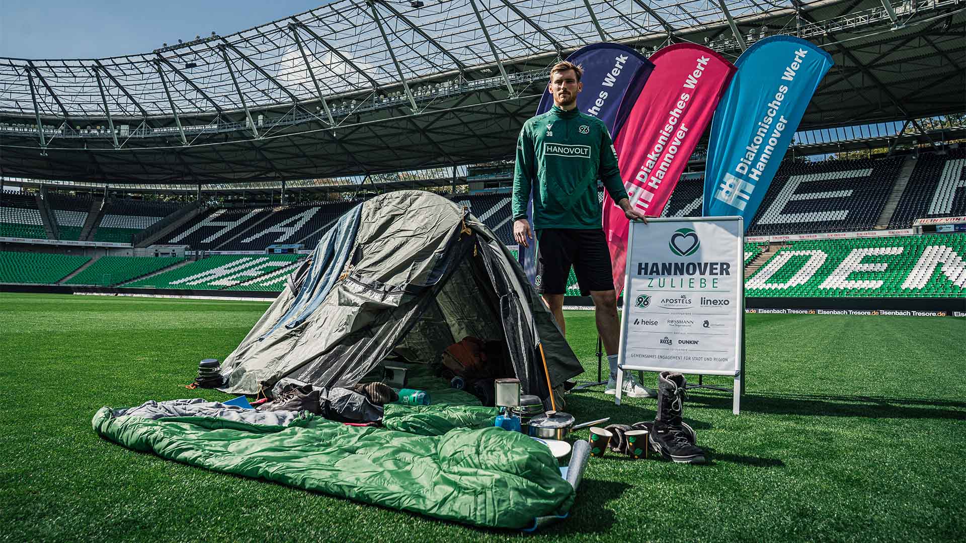 Der 96-Torhüter Leo Weinkauf steht in Trainingskleidung im 96-Stadion, neben ihm ist ein kleines Zelt aufgebaut, hinter ihm stehen Beachflags vom Diakonischen Werk, er legt die Hand auf ein Schild mit der Aufschrift HANNOVER ZULIEBE und Partnerlogos. 
