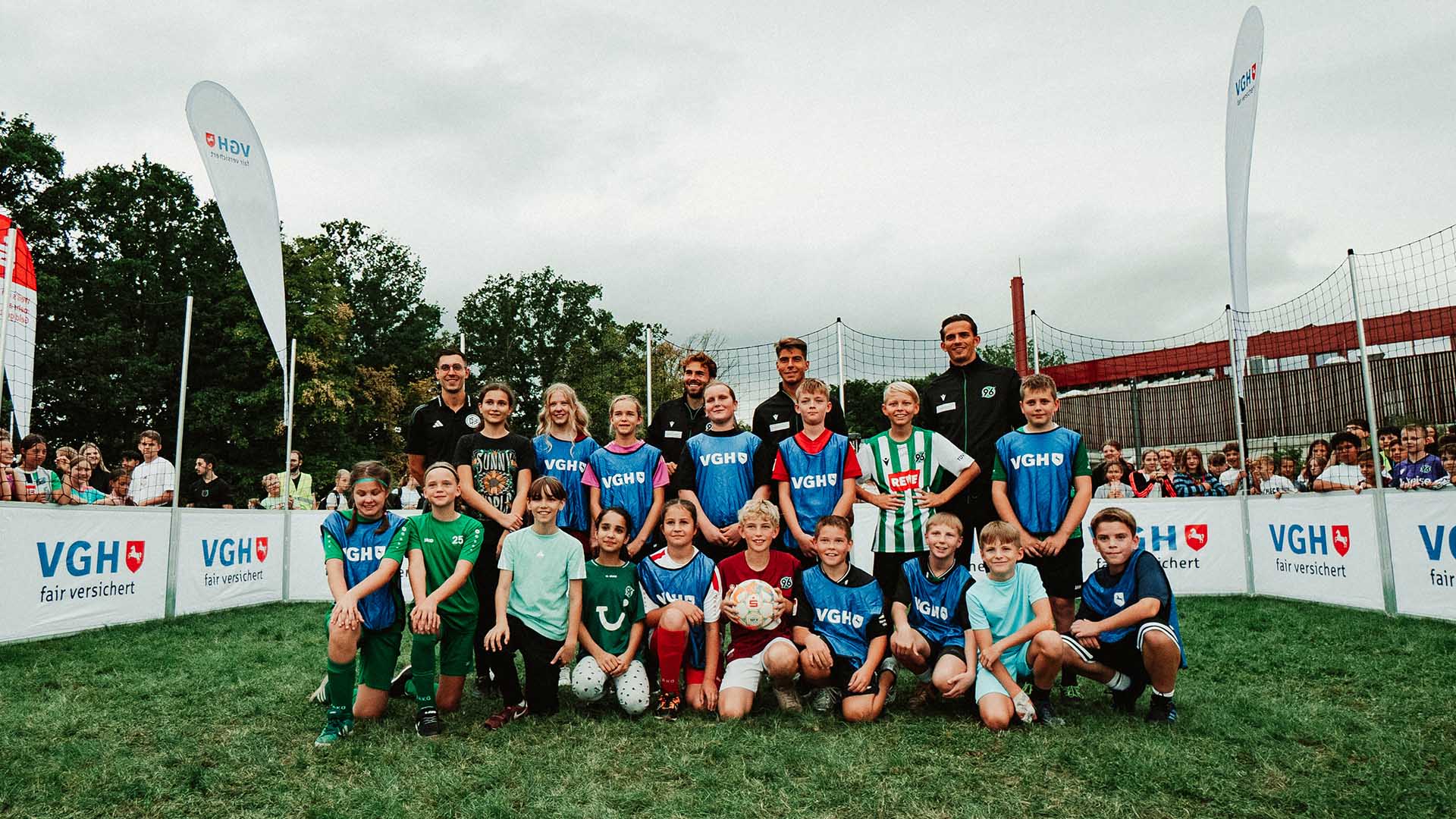 Eine Gruppe Kinder posiert lächelnd mit drei Fußball-Profis und einem Schiedsrichter für ein Bild.