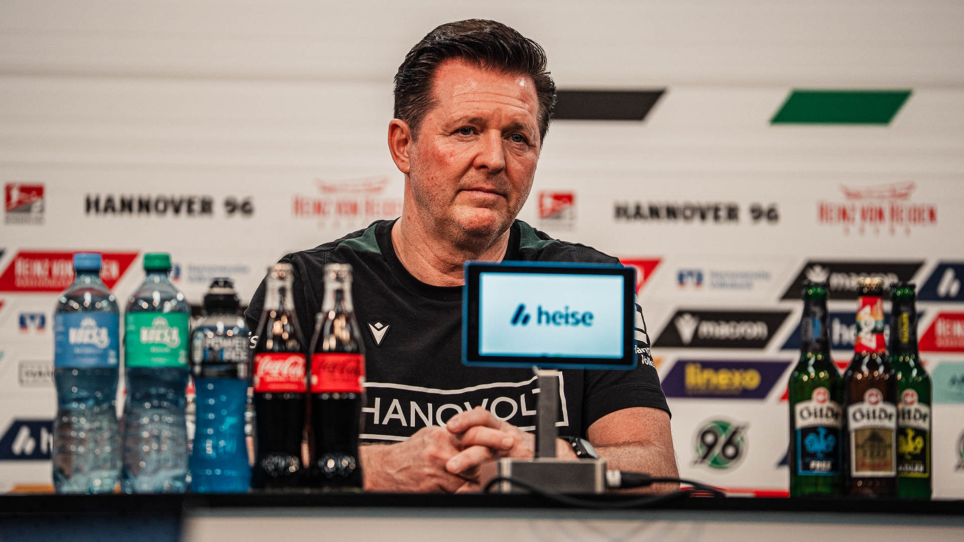 96-Cheftrainer Christian Titz sitzt auf dem Pressekonferenz-Podium, hinter ihm eine Sponsorenwand, vor ihm ein Mikro und verschiedene Getränkeflaschen.
