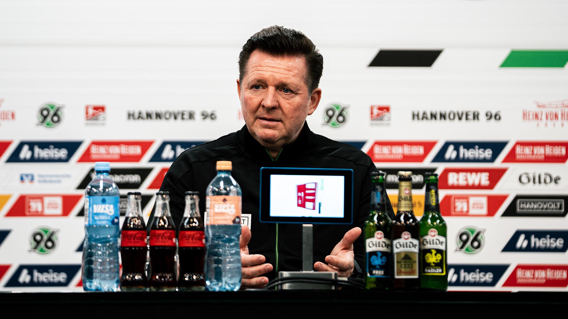Christian Titz sitzt auf dem PK-Podium, vor ihm Getränkeflaschen und das Mikro, dahinter die Sponsorenwand.