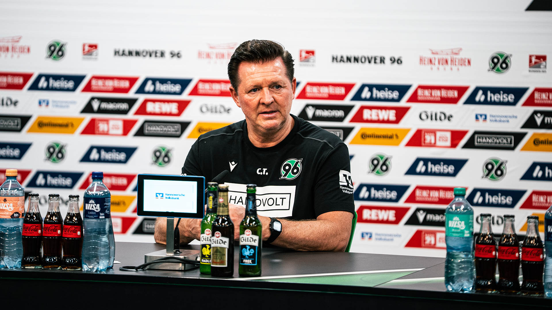 Christian Titz sitzt im schwarzen 96-T-Shirt auf dem PK-Podium.