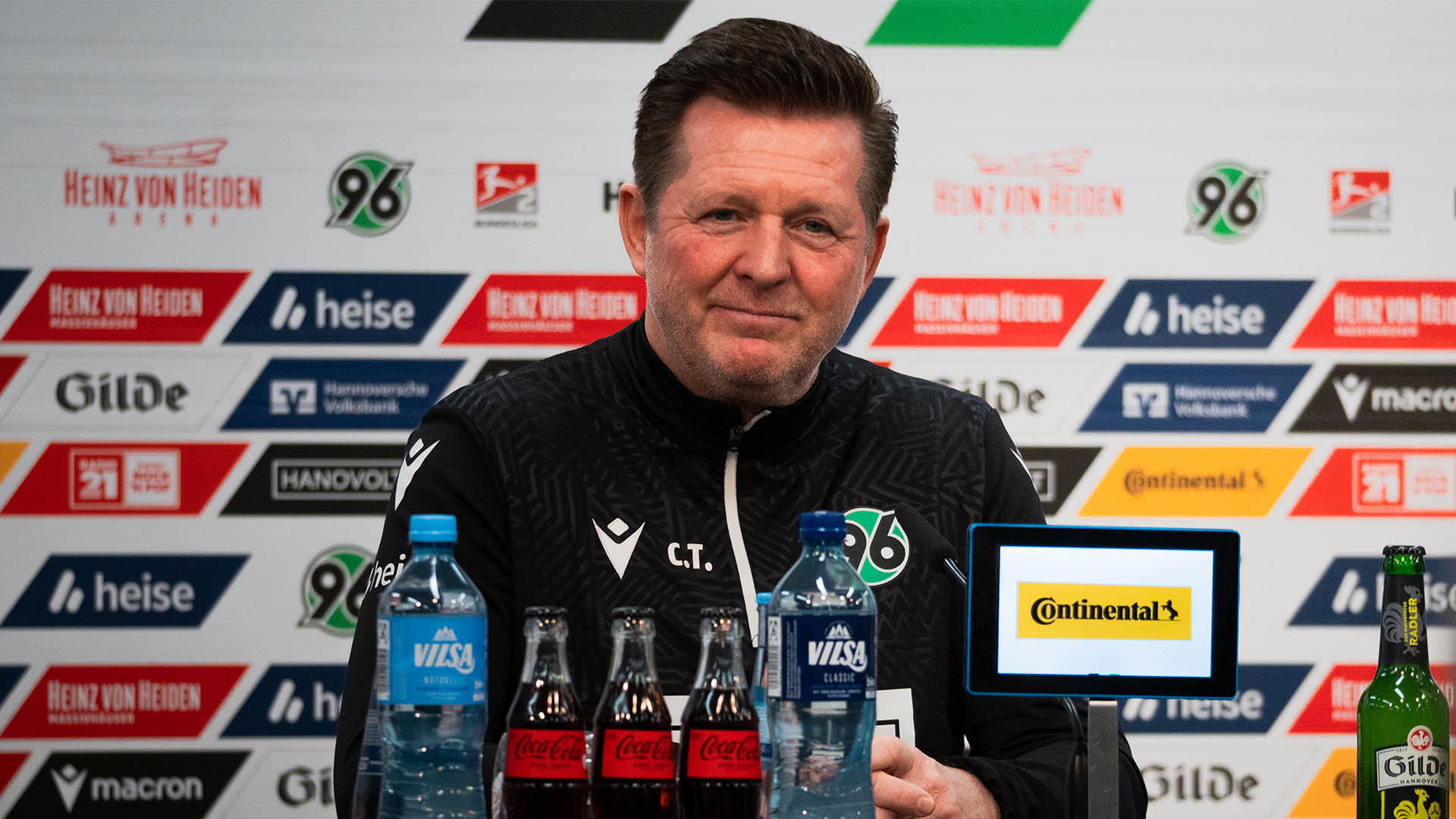 Christian Titz sitzt leicht lächelnd auf dem Podium, hinter ihm eine Sponsorenwand, davor Flaschen und Sponsoren-Tablets.