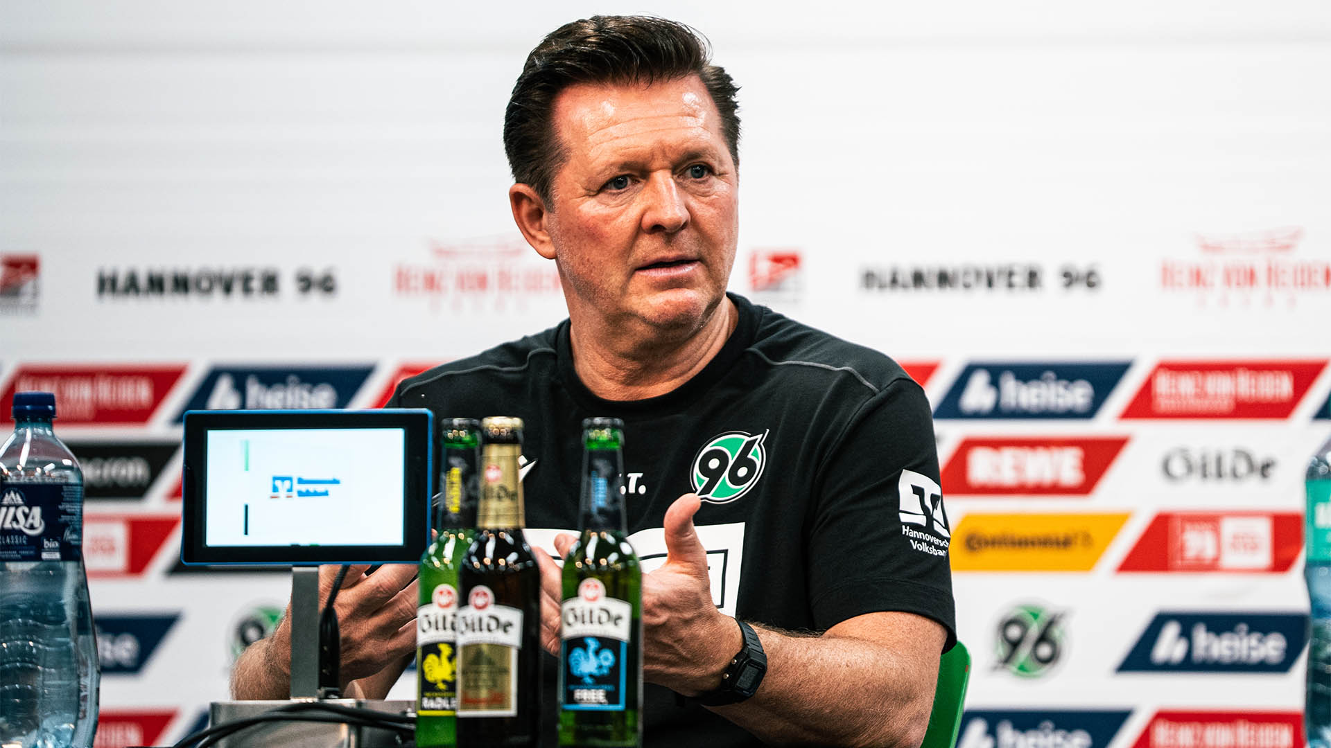 Christian Titz sitzt auf dem PK-Podium, hinter ihm die Sponsorenwand, vor ihm ein Werbetablet und Gildeflaschen.