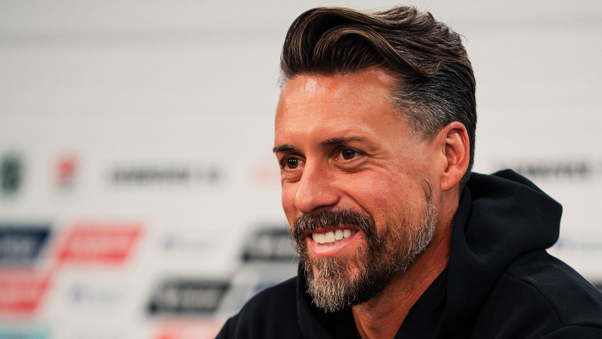 Der neue 96-Trainer sitzt im schwarzen 96-Hoodie auf dem Pressekonferenz-Podium, er lacht freundlich.