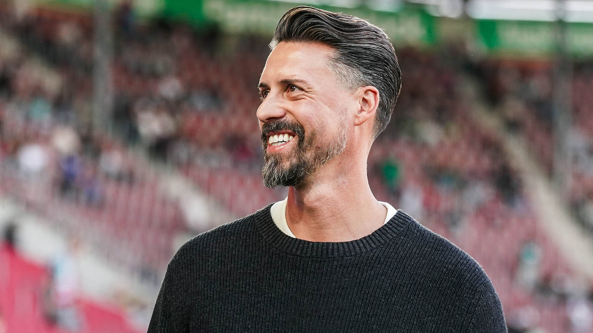 Der neue Trainer Sandro Wagner ist seitlich zu sehen, er trägt einen dunklen Wollpullover und lacht herzlich.