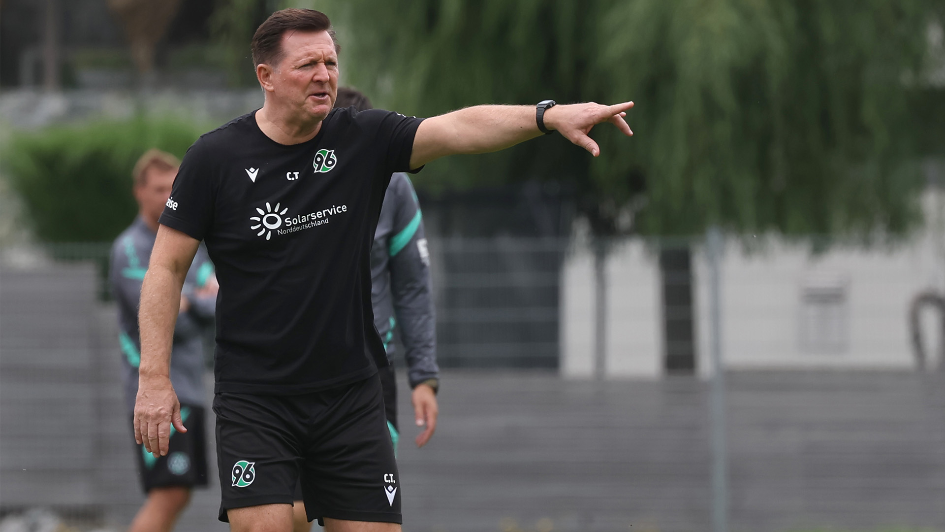 Hannover-96-Trainer Christian Titz steht auf dem Platz und zeigt mit dem ausgestreckten linken Arm nach links.