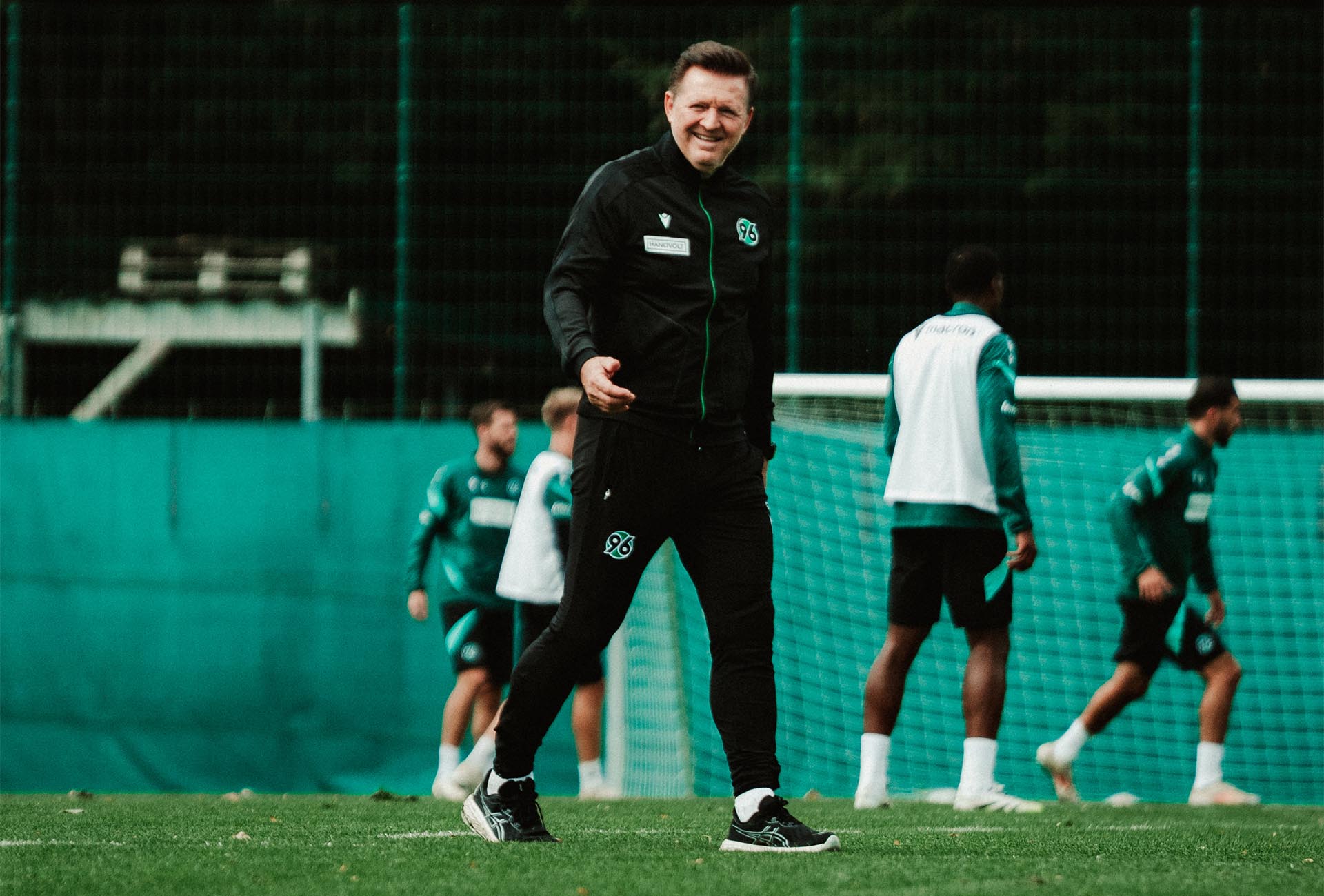 Christian Titz läuft lächelnd über den Trainingsplatz, im Hintergrund unscharf einige Spieler.