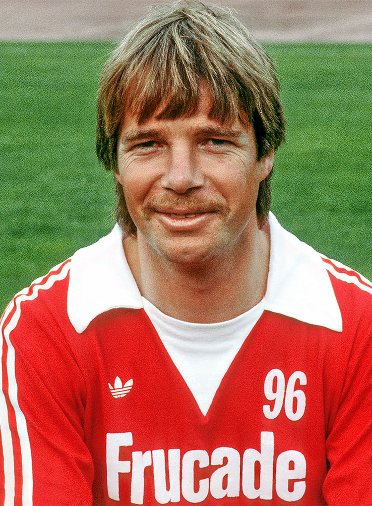 Ein Mann im roten 96-Trikot lächelt in die Kamera, im Hintergrund grüner Rasen.