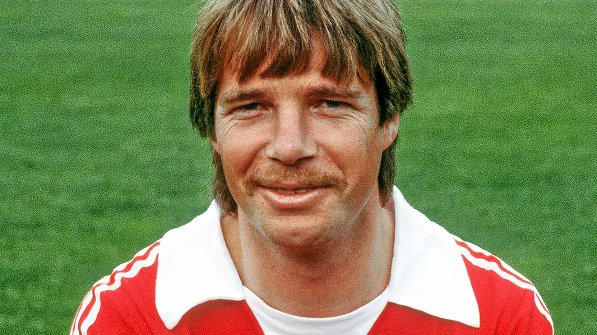 Ein Mann im roten 96-Trikot lächelt in die Kamera, im Hintergrund grüner Rasen.