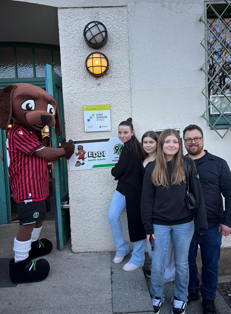 Das KIDS-Maskottchen EDDI und eine Gruppe von vier Personen stehen vor der weißen Wand, an der die Plakette angebracht wurde.