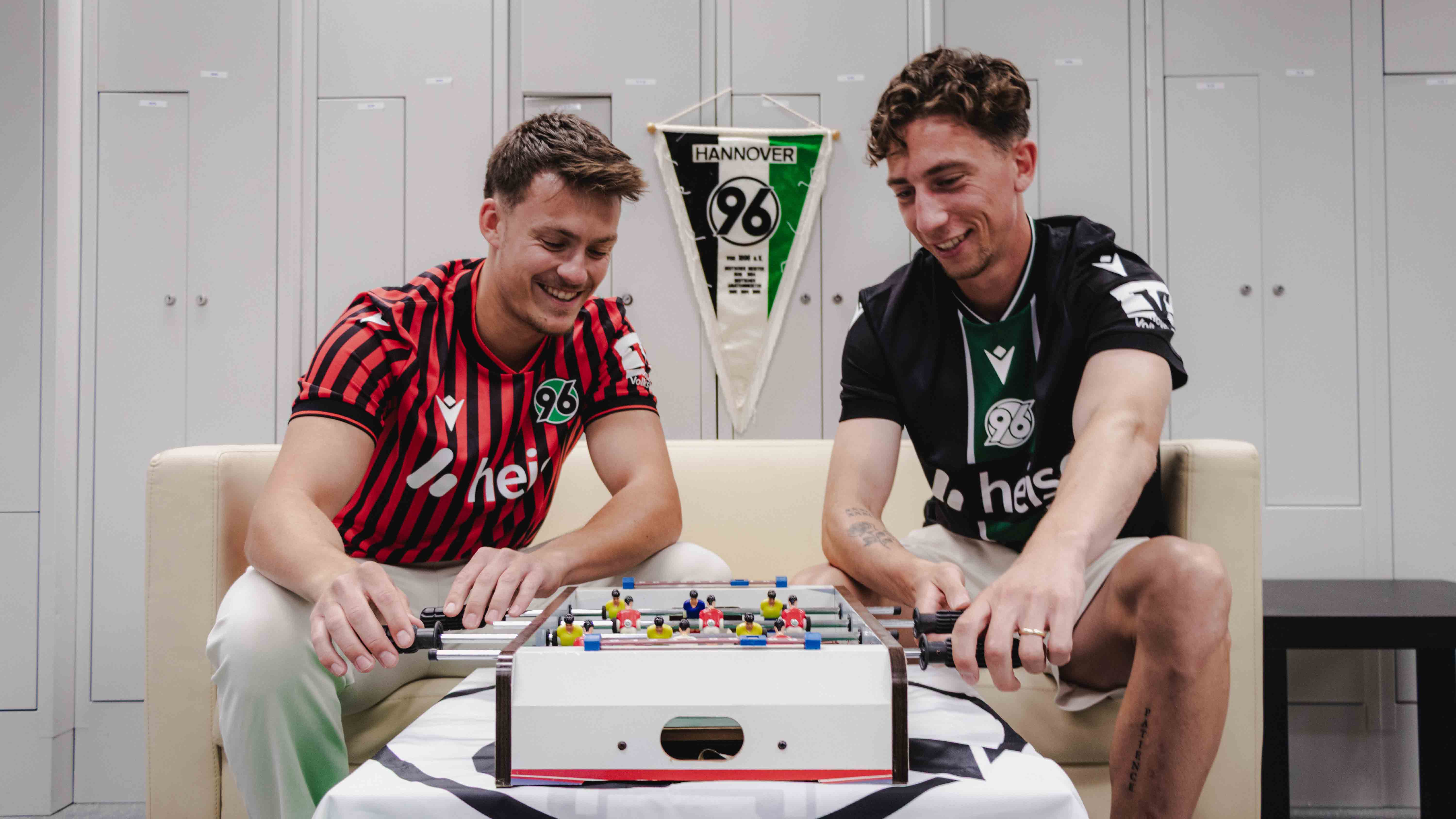 Host Niklas (links) und Rochelt sitzen auf dem Sofa und spielen Tischfußball.